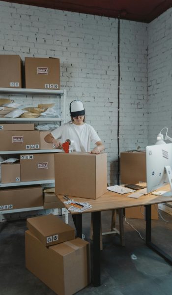 A warehouse worker in uniform packaging boxes for delivery in an organized storage area.